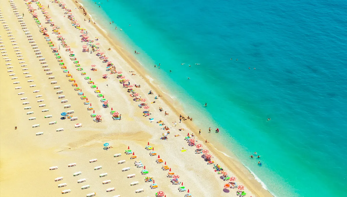 Aerial view of a crowded sandy beach with colorful umbrellas and people swimming in the turquoise sea.