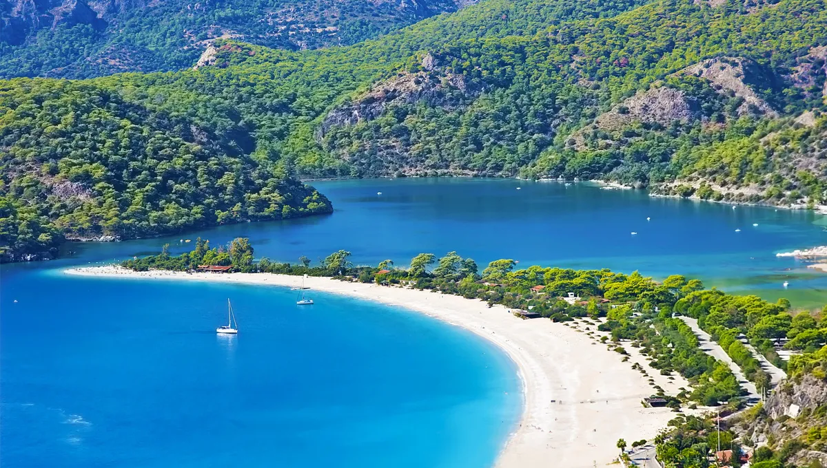 Olu Deniz beach in Turkey with clear blue water, sandy shore, and green forested hills surrounding the bay.