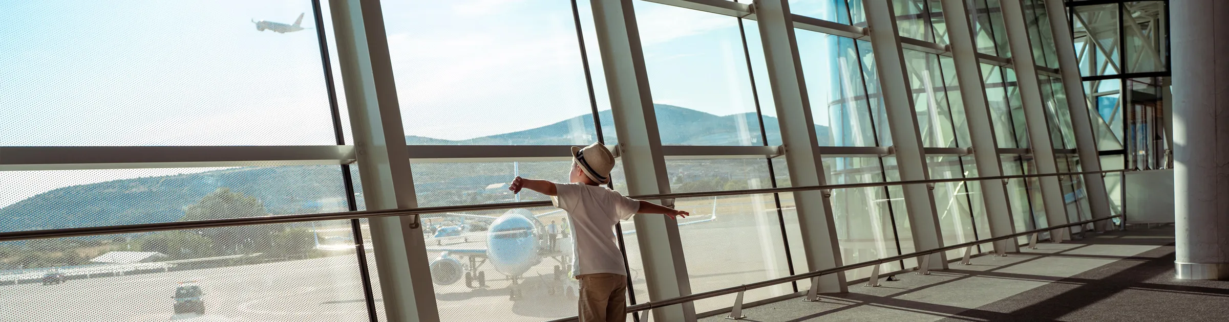 Child watching airplanes through large airport terminal windows with mountains in the background.