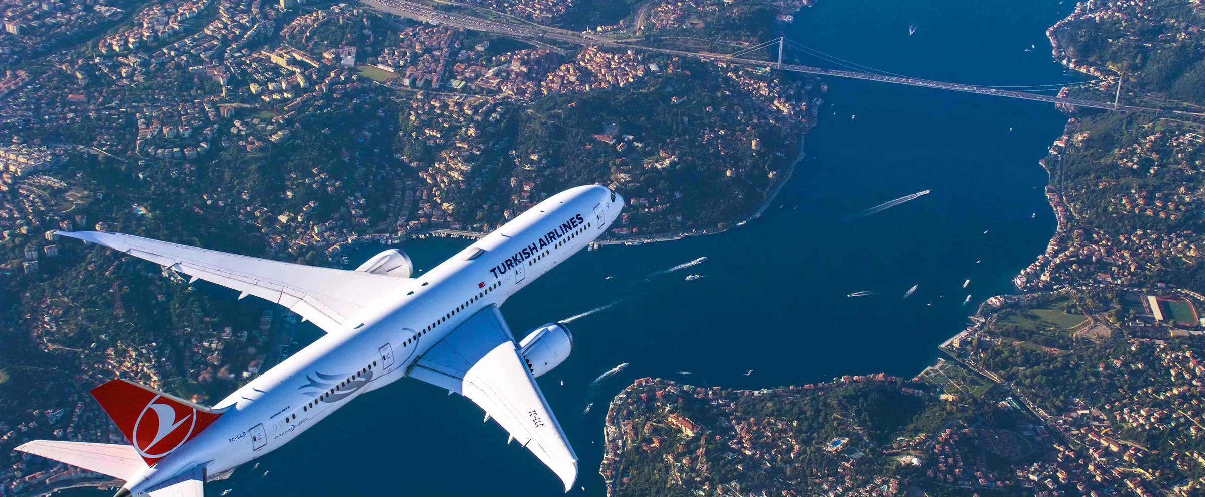 Commercial airplane flying over Istanbul with the Bosphorus Bridge and sea traffic visible below the plane.