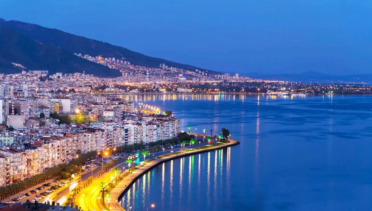 Vue nocturne d'Izmir avec des lumières côtières se reflétant sur l'eau et des montagnes au loin.