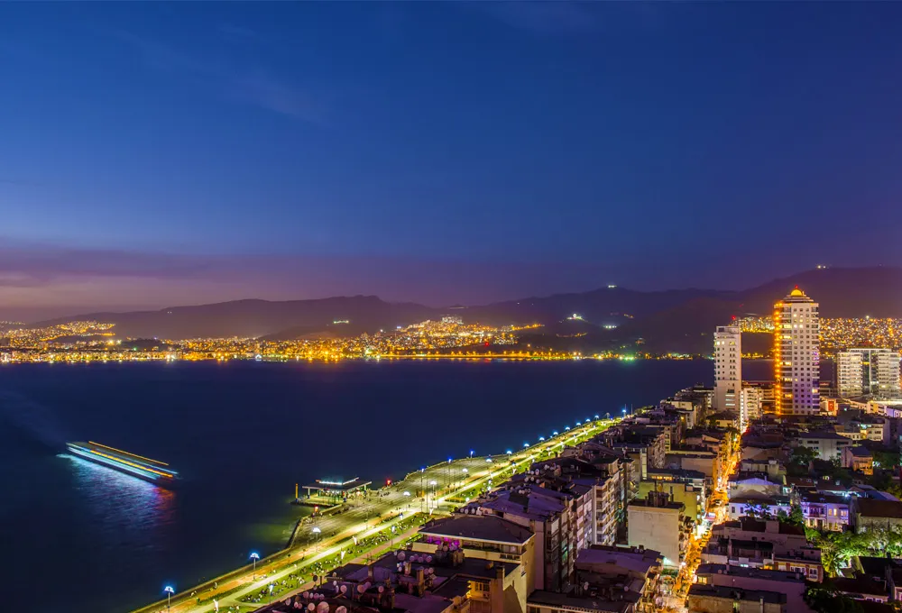Vue nocturne de la côte d'Izmir avec des bâtiments illuminés et un bateau naviguant, location de voitures en Turquie.