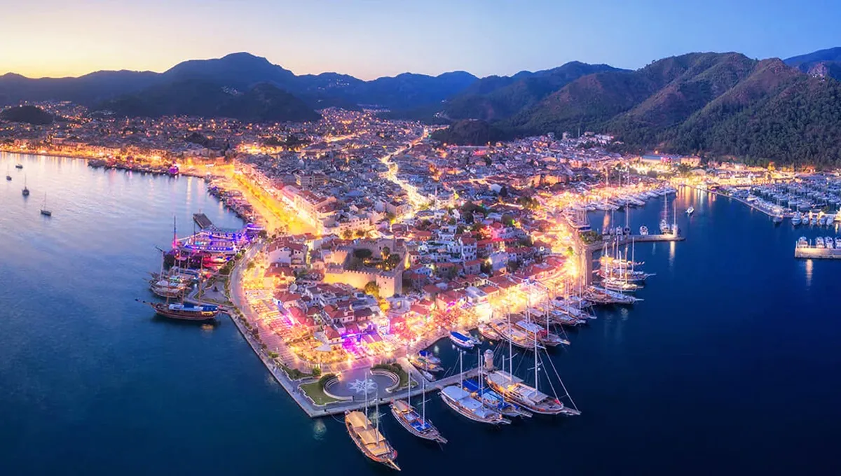 Marmaris sahilinde gün batımında ışıklı sokaklar ve limanda demirlemiş tekneler araba kiralamaya uygun.