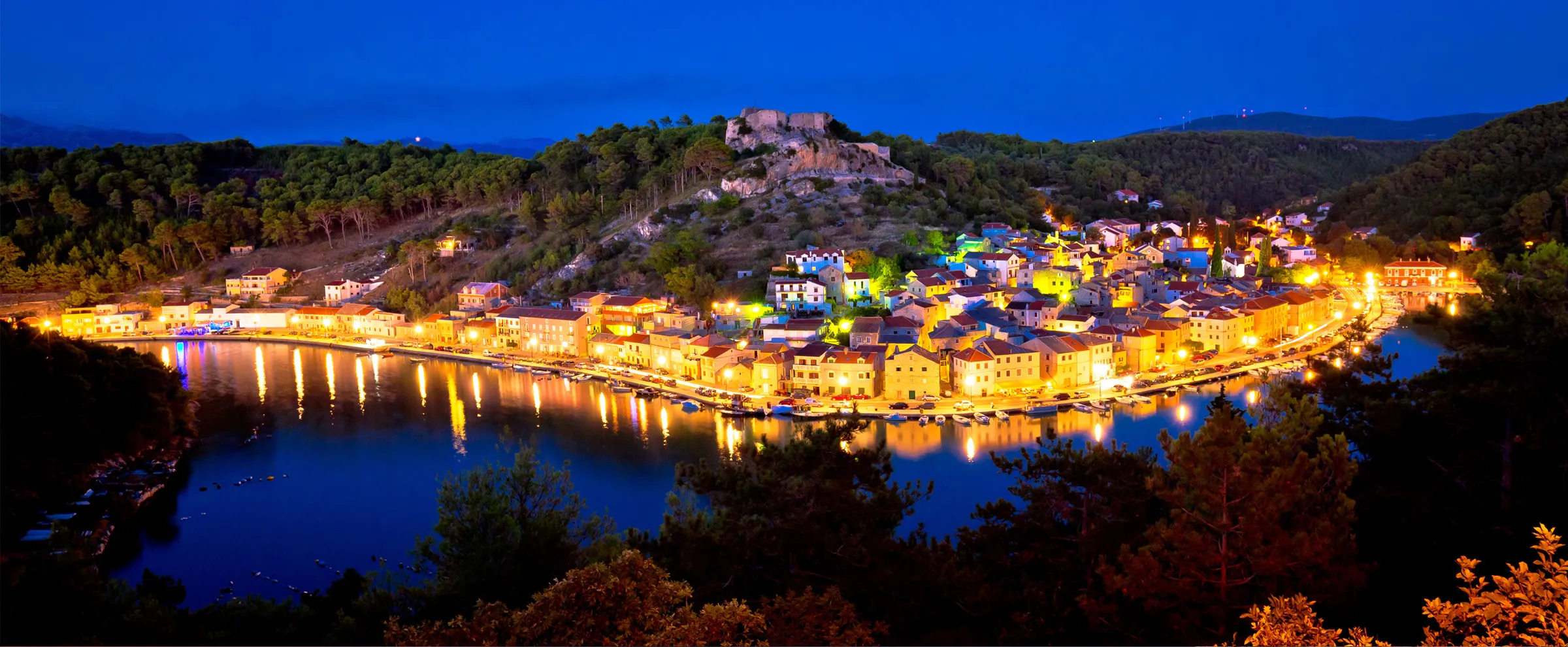 Night view of Cres town bay with illuminated buildings reflected on calm water in Croatia.