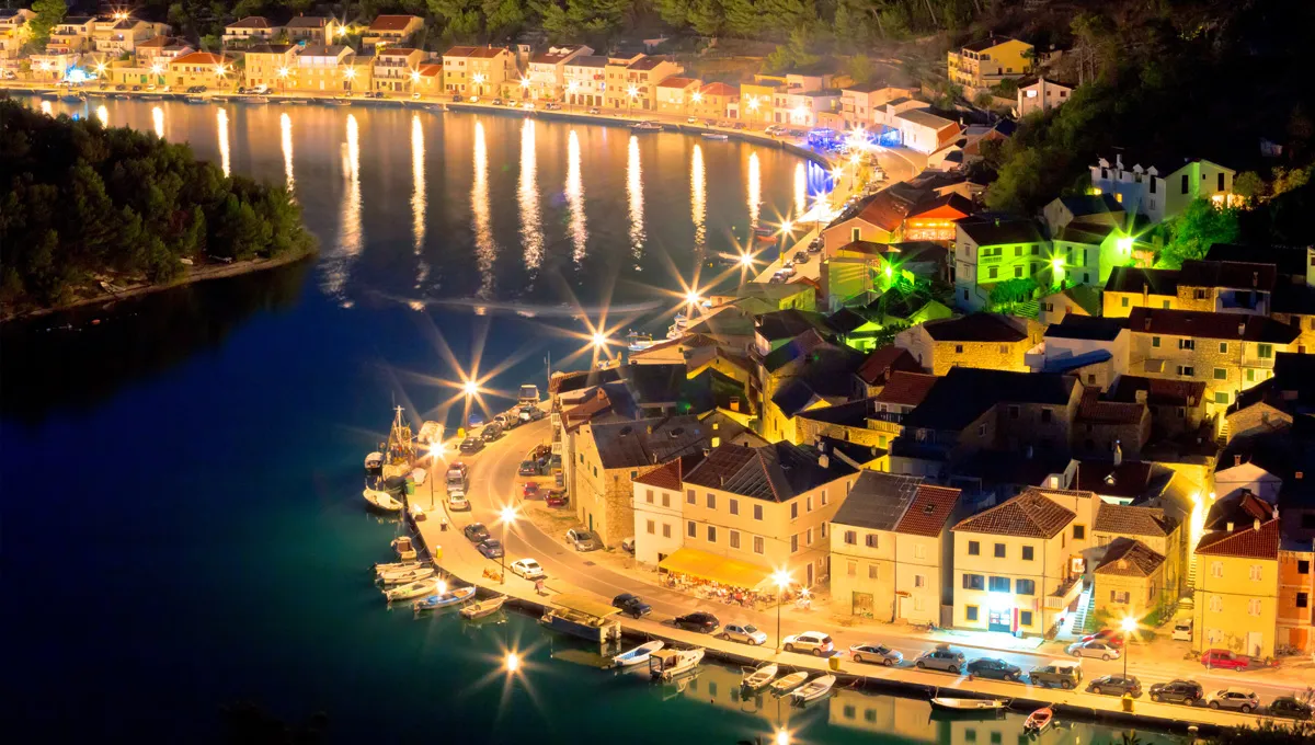 Night view of a coastal town with illuminated streets and waterfront parking for car rental.