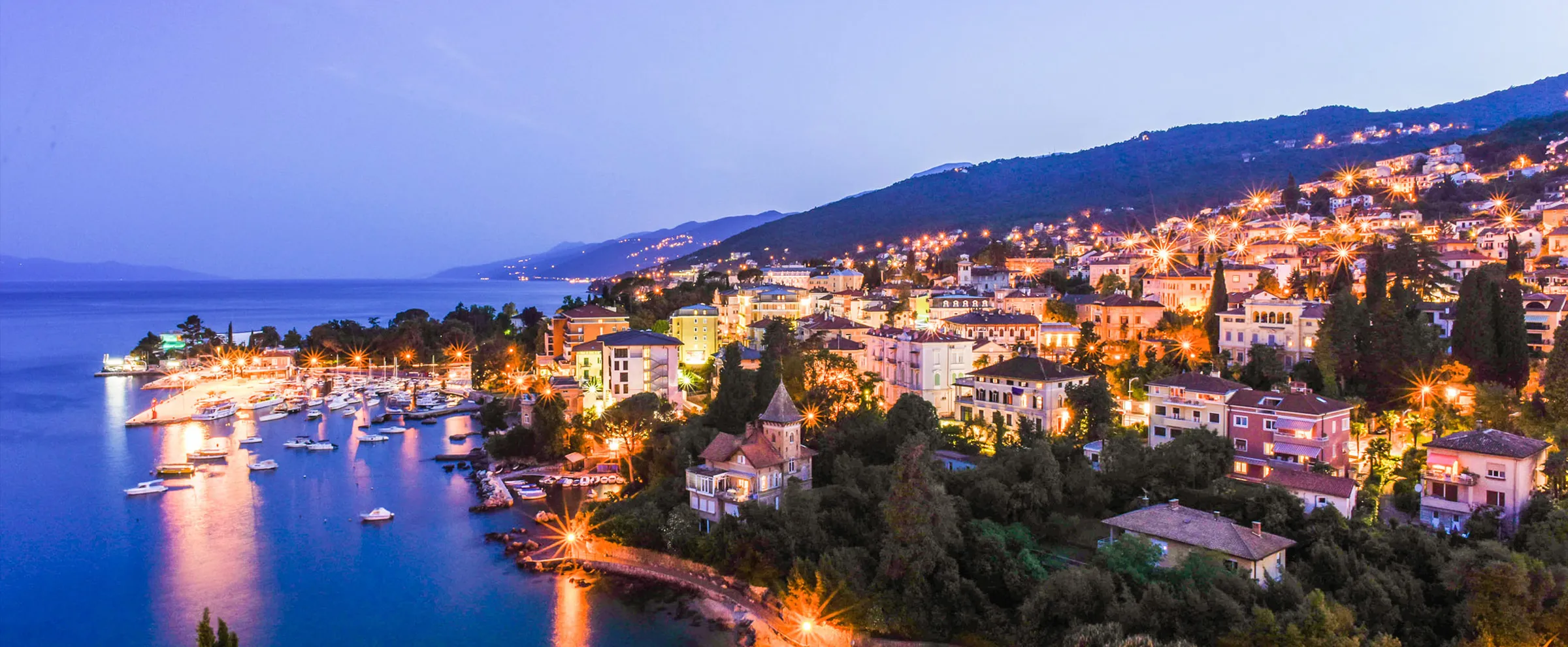 Opatija coastline at twilight with illuminated buildings and boats in the bay, ideal for car rental trips.