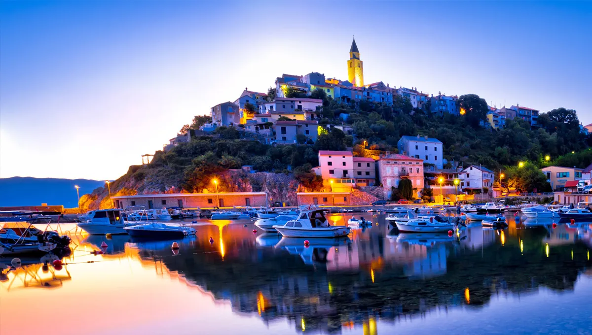 Mošćenička Draga harbor at dusk with boats docked and illuminated houses on the hillside.