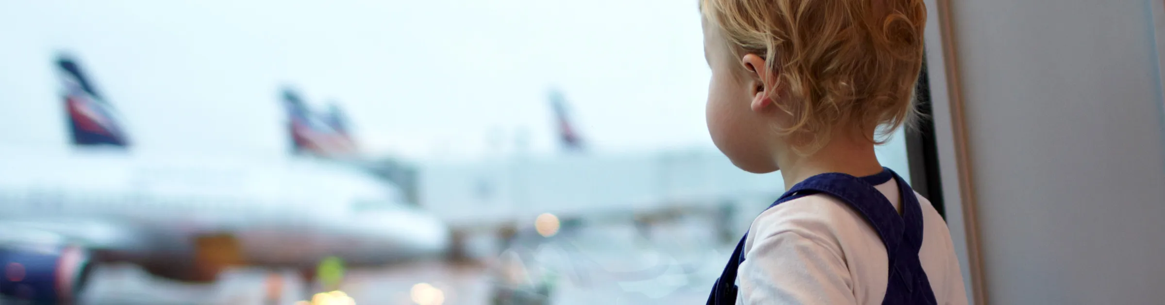 Child looking out airport window at planes on the runway, suitable for car rental airport transfer service.