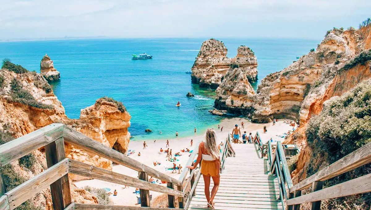 Lagos beach in Portugal with bright cliffs, turquoise sea, and a woman walking down wooden stairs to the sand.