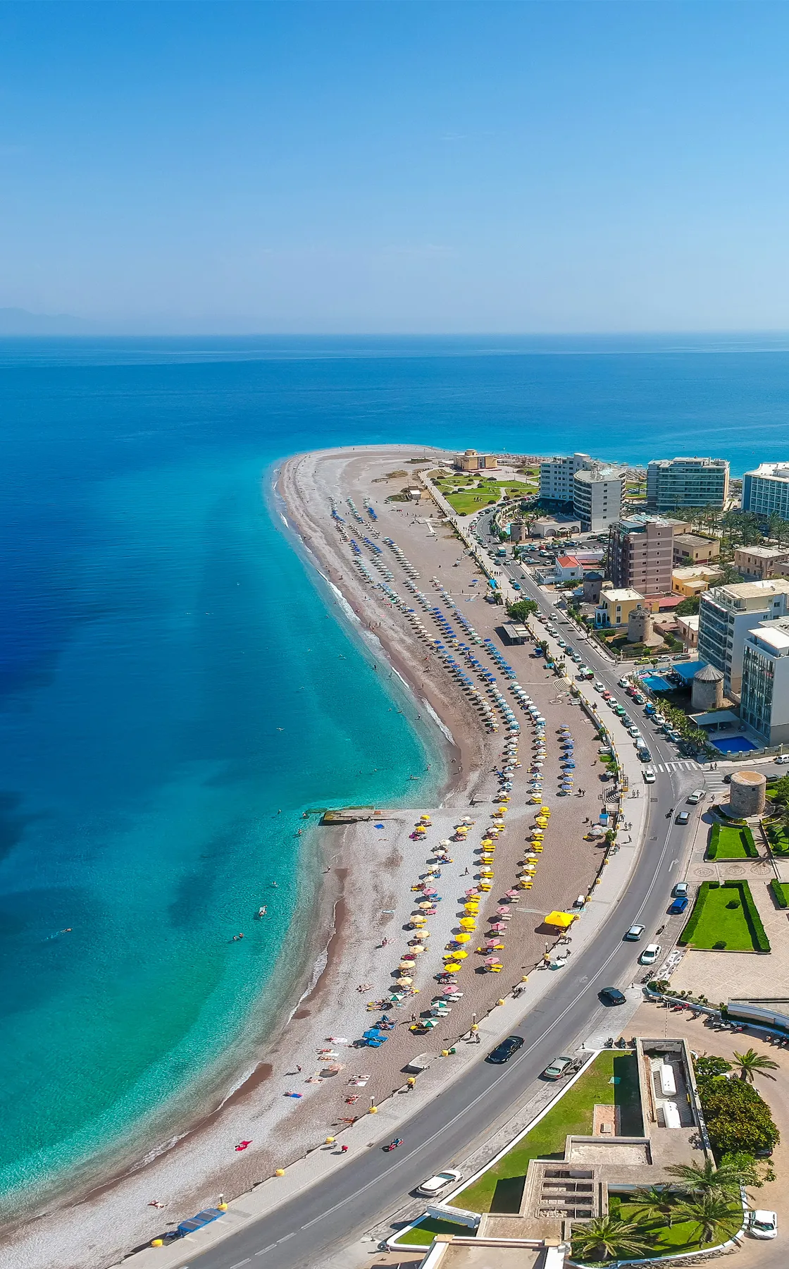 Rhodes beach with colorful umbrellas and clear blue sea along a coastal road with buildings and cars.