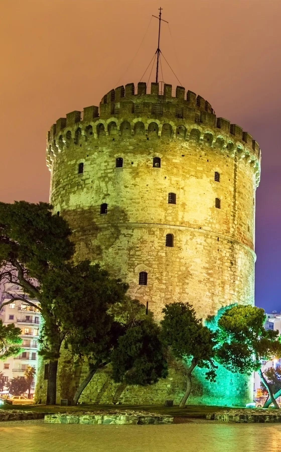 White Tower of Thessaloniki lit at night with surrounding trees and nearby buildings visible.