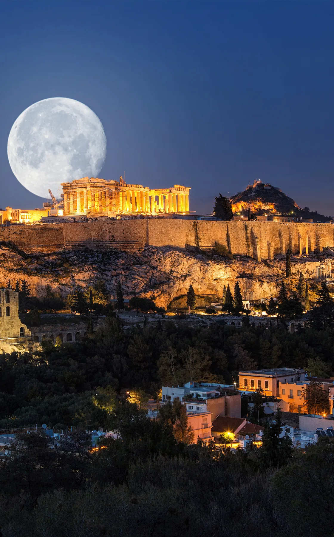 Athens Acropolis illuminated at night with a large full moon in the sky above the ancient ruins.