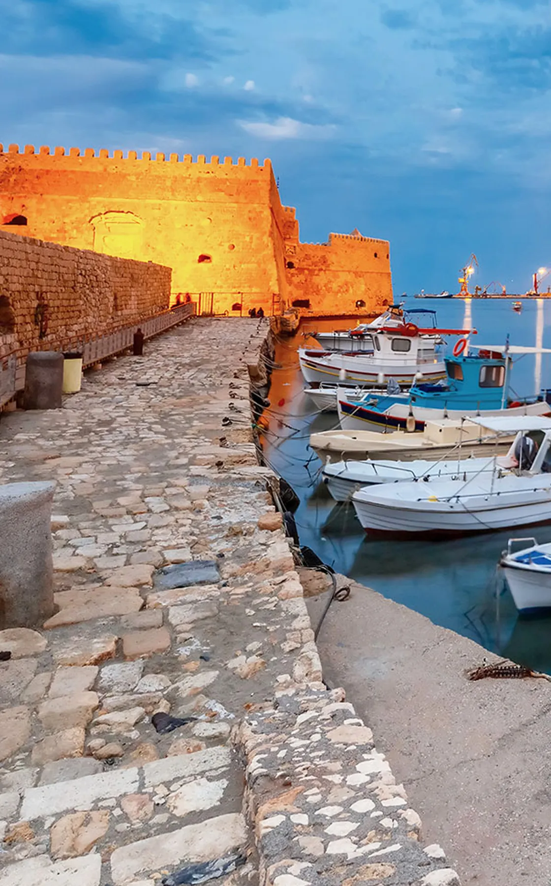Heraklion harbor with small boats docked by a stone pier near a historic fortress at sunset.