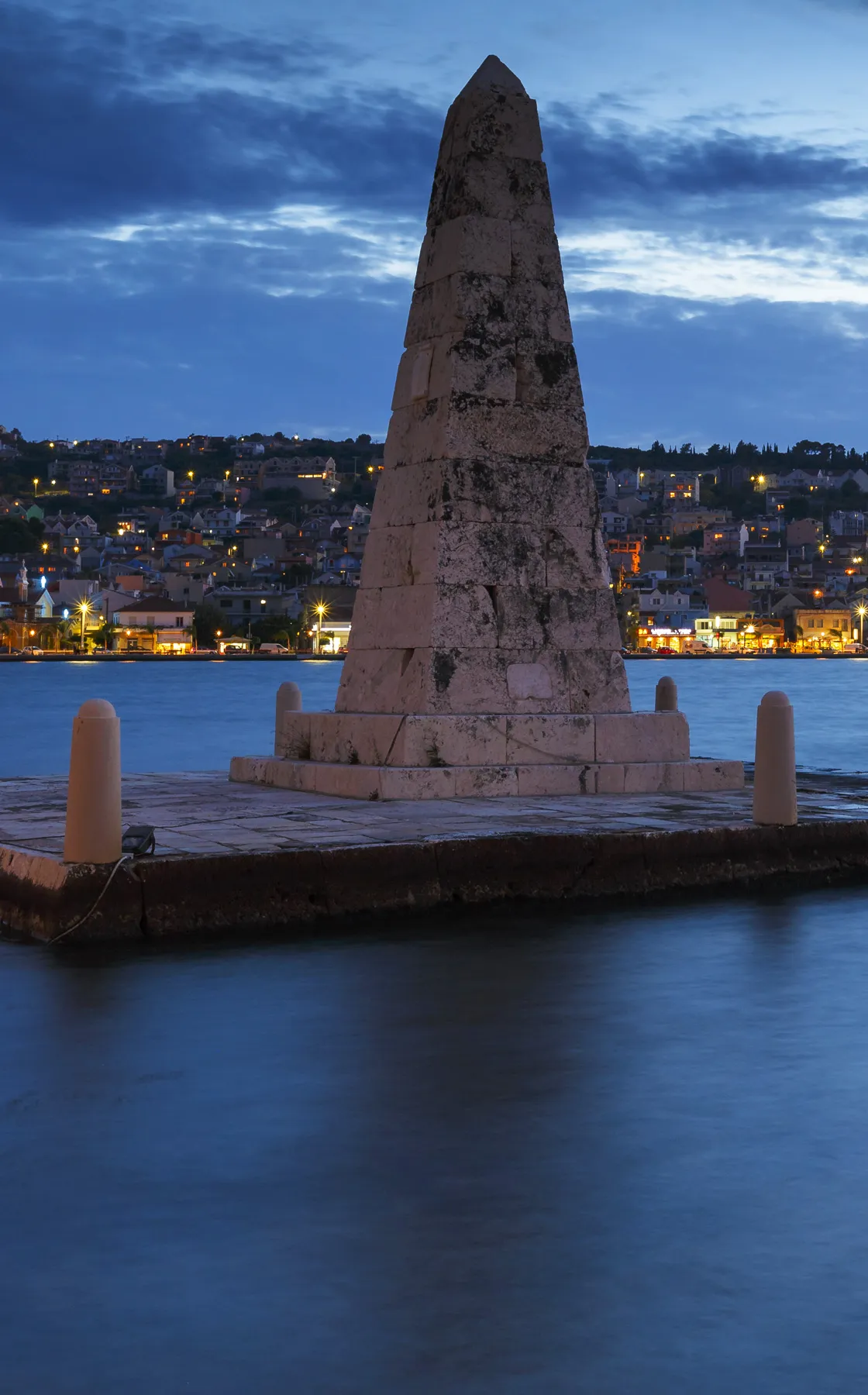 Stone obelisk monument by the sea with coastal town lights in the background at dusk for car rental trips.