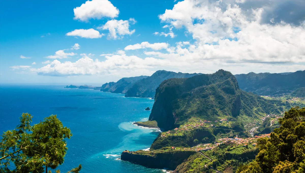 Costa da Madeira com falésias, colinas verdes e oceano azul sob um céu parcialmente nublado para aluguer de carros.