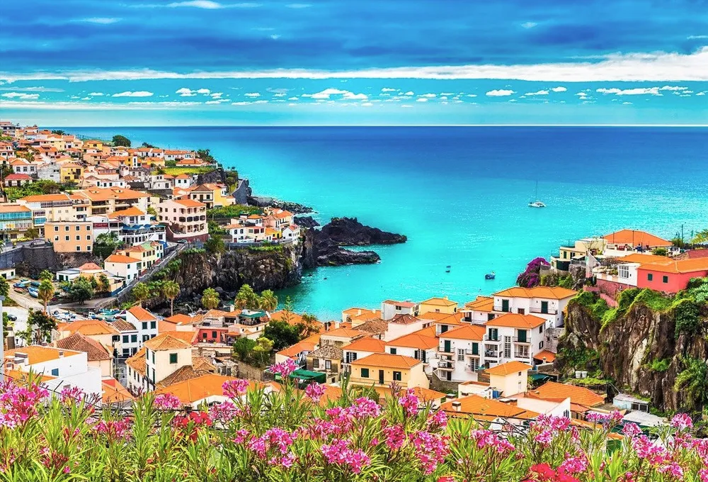 Costa da Madeira com casas brilhantes, mar turquesa e flores, perfeita para aluguer de carros junto ao mar.