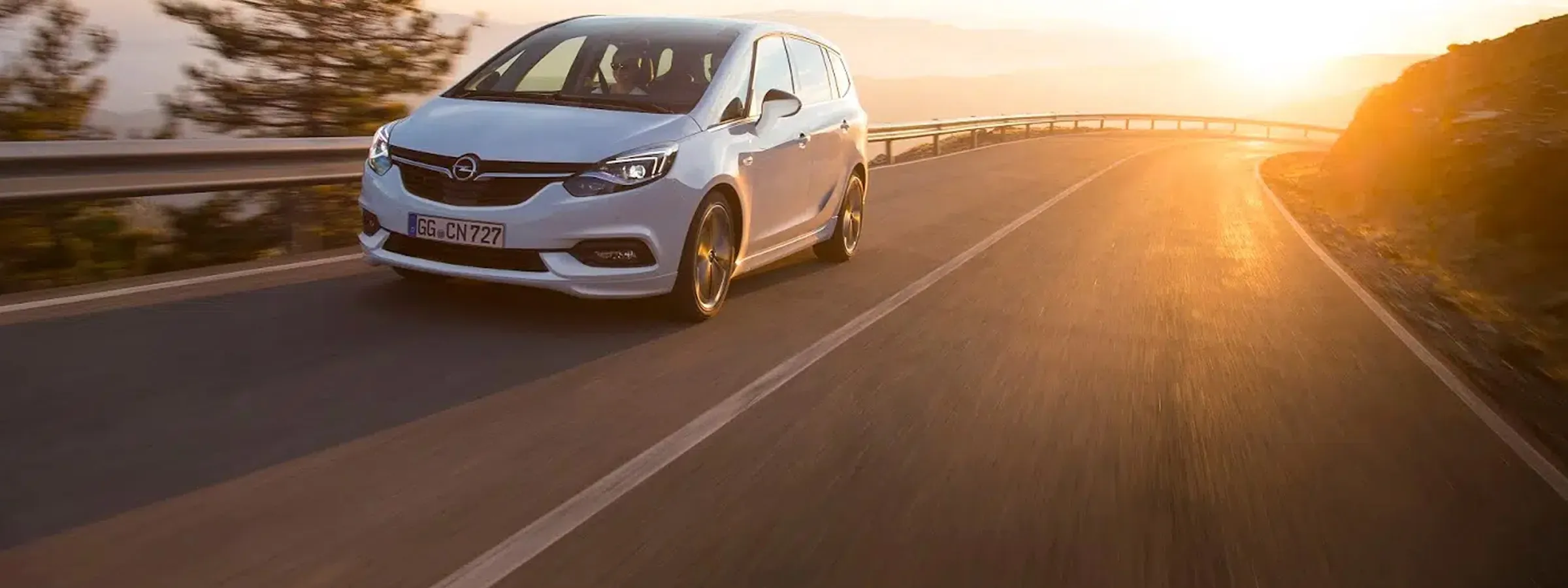Opel car rental driving on a coastal road at sunset with bright sun and clear sky.