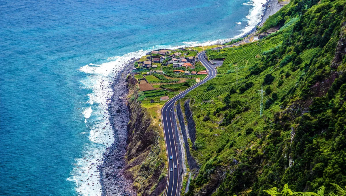 Coastal road with cars driving along a cliff by the ocean near a small village and green fields.