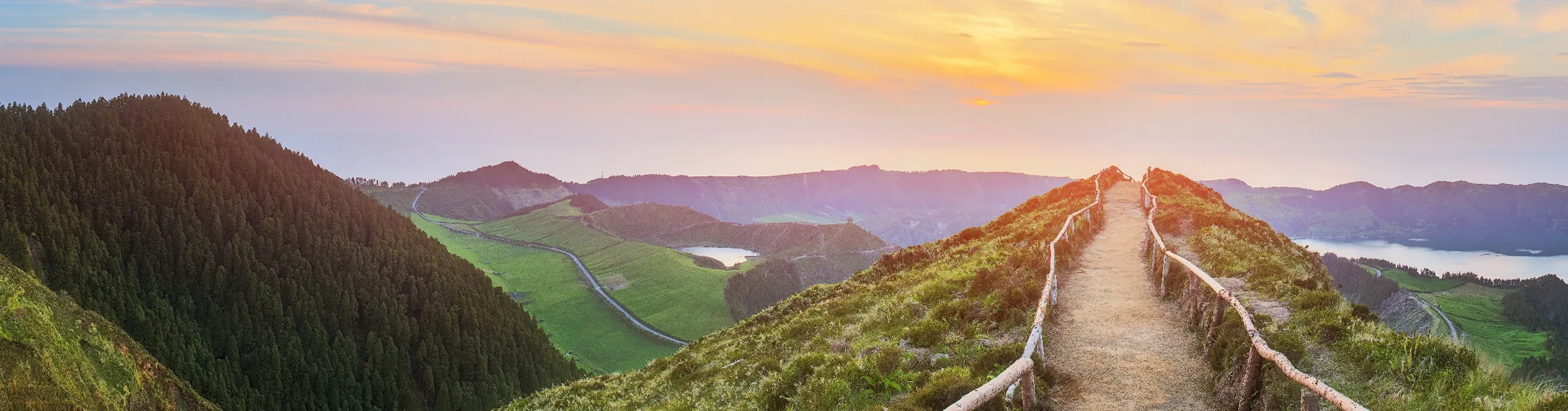 Sunset over a scenic mountain path surrounded by lush greenery and distant lakes.