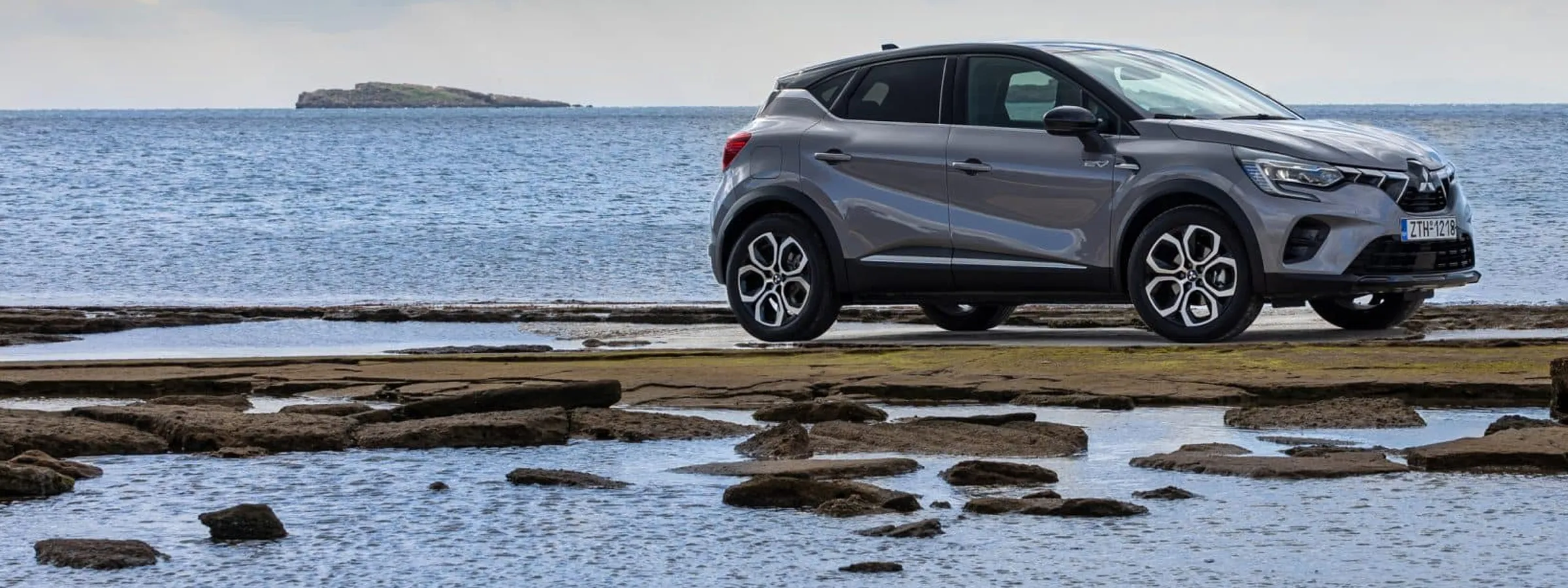 Renault Captur car rental parked by the rocky seashore with calm sea and small island in the distance.