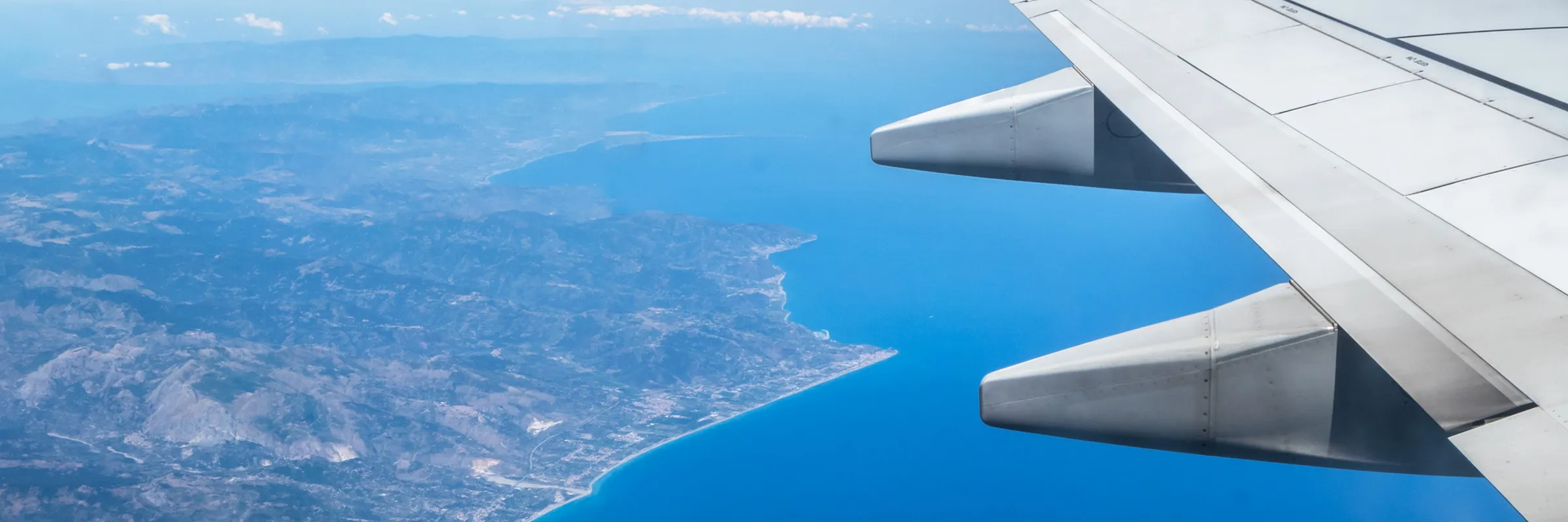 Airplane wing flying over a coastal area with blue sea and mountainous land below seen from above.