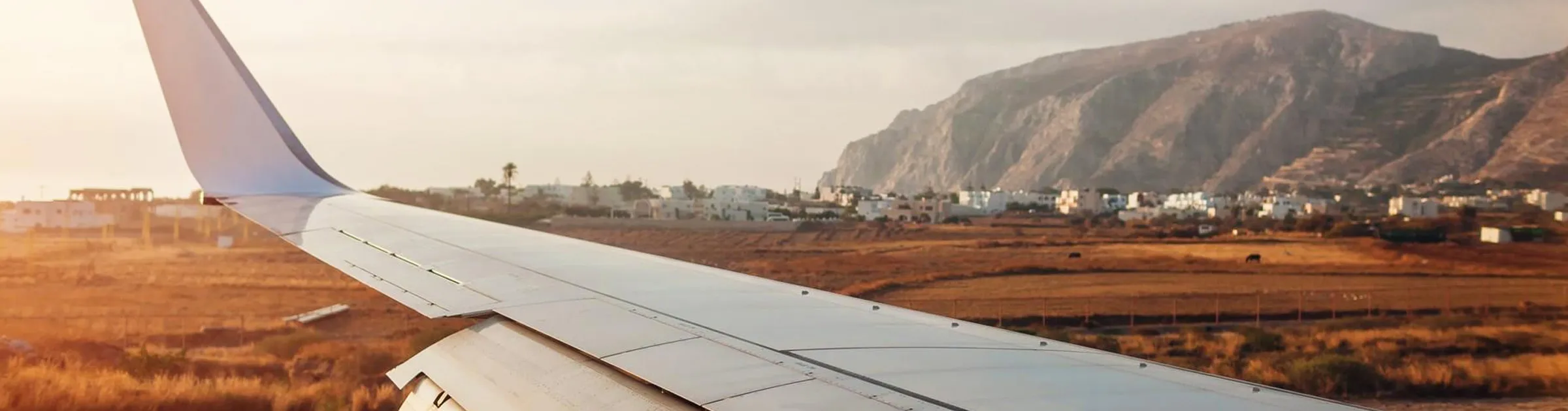 Aile d'avion au-dessus d'un paysage sec avec des bâtiments et des montagnes à distance au coucher du soleil.