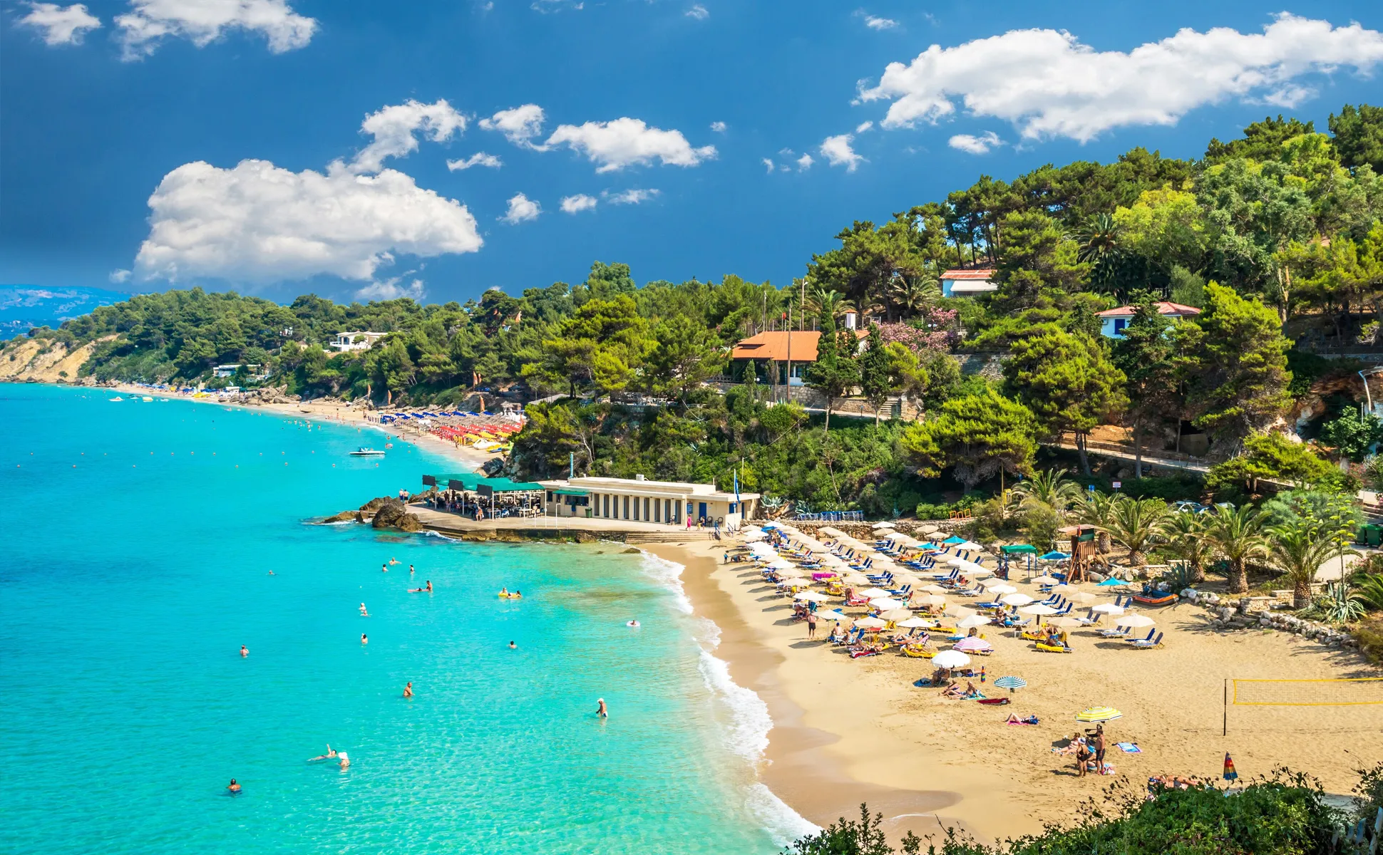 Saint Thomas Beach in Skopelos with sunbathers, umbrellas, turquoise sea, and green pine-covered hills nearby.