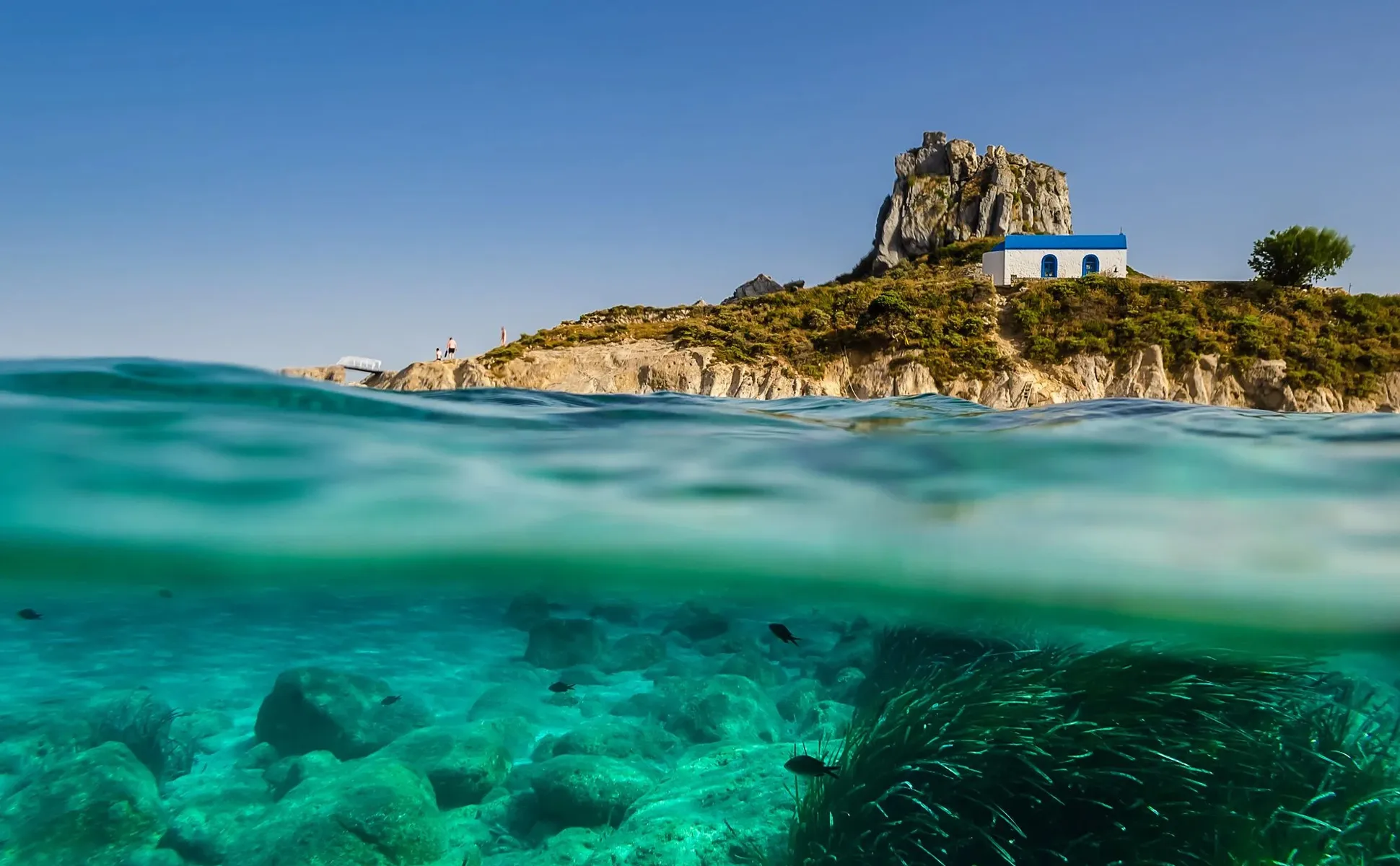 Coastal view of a small white chapel with blue roof on rocky land by clear turquoise sea water.