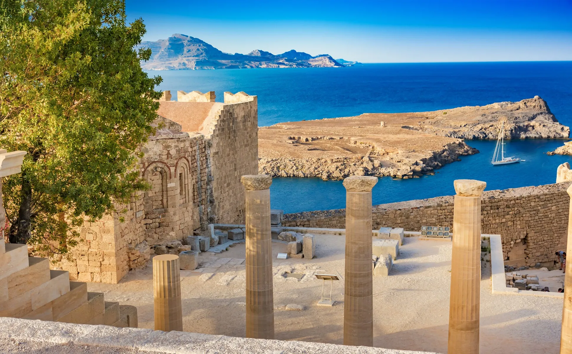 Ancient ruins with stone columns and a boat on the blue sea near a rocky coastline.