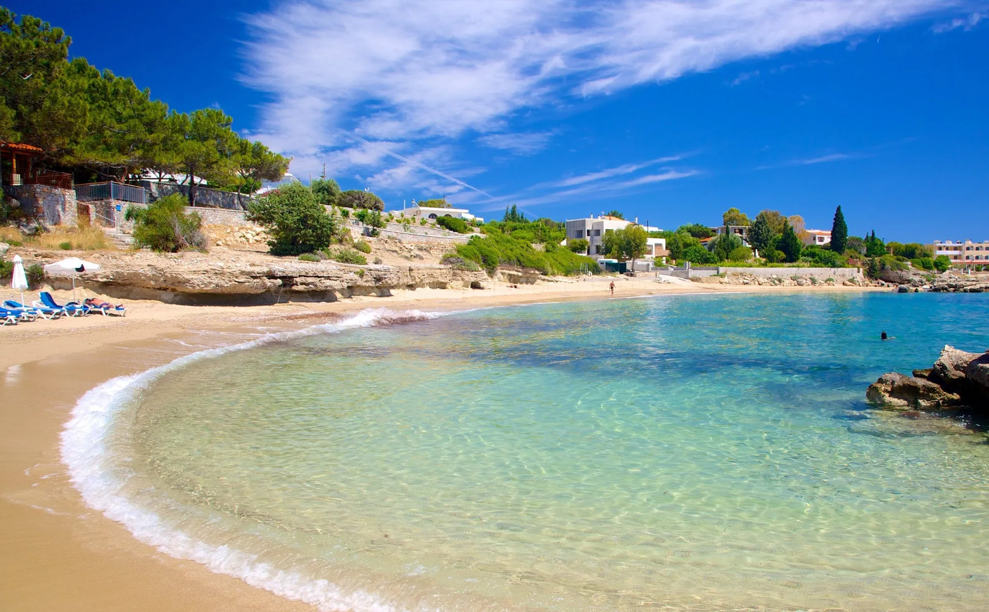 Clear water shoreline and sandy beach under blue skies with scattered clouds and nearby houses.