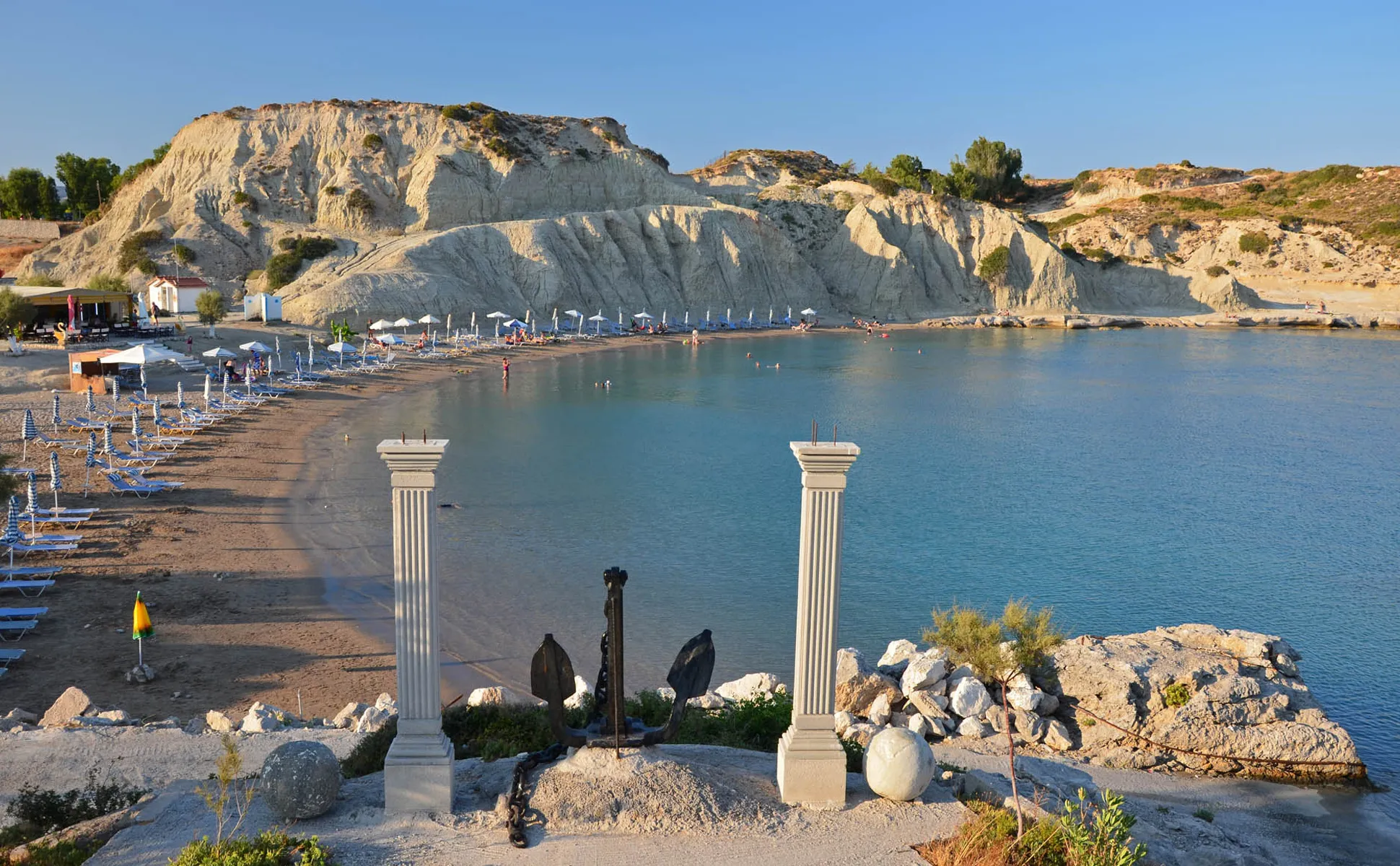 Plakias Beach in Crete with sandy shore, sun loungers, and rocky cliffs by calm blue sea water.