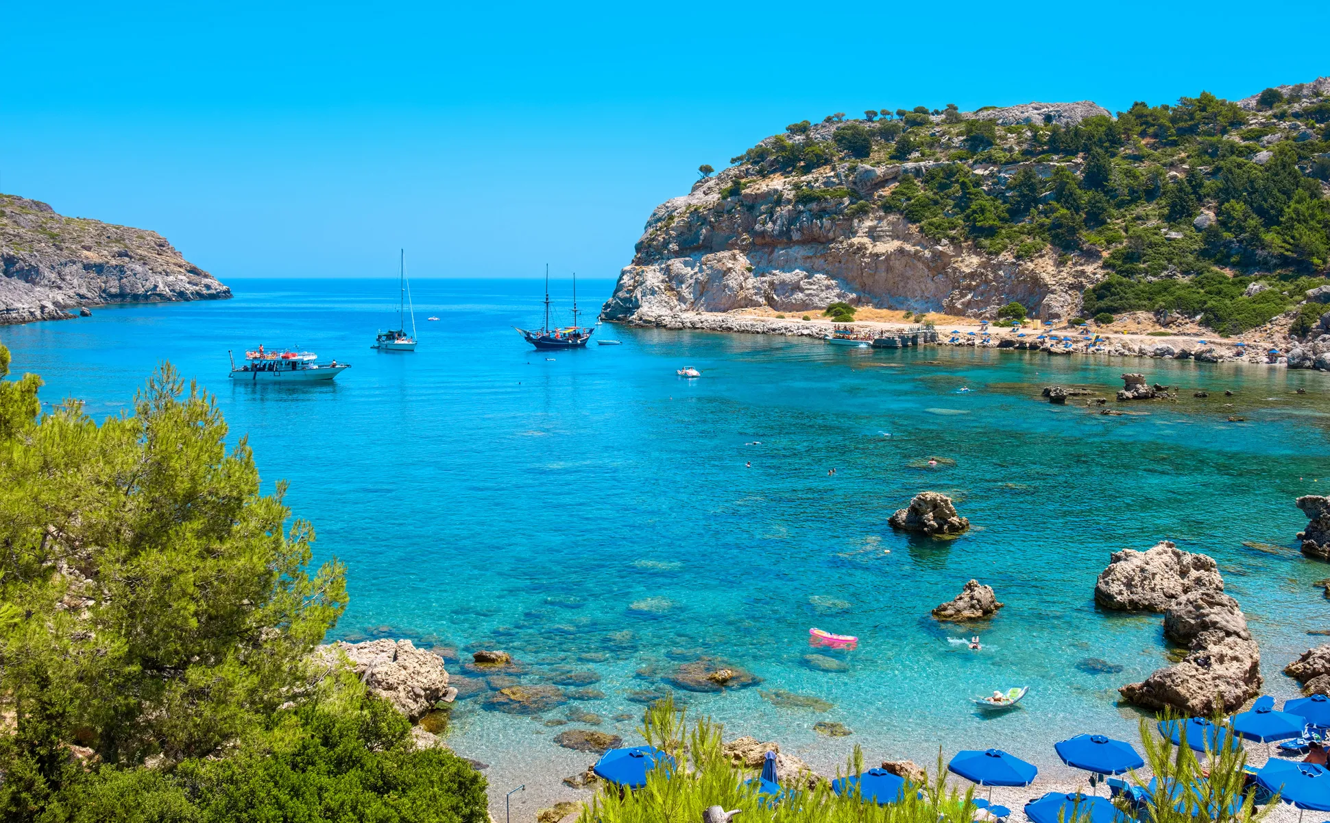 Rhodes beach cove with blue sea, boats anchored, rocky cliffs, and sun umbrellas along the shore.