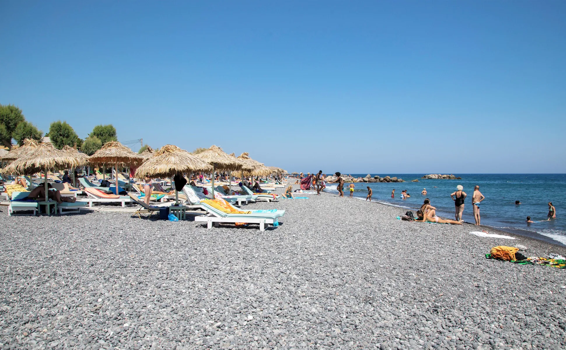 Pebble beach with sunbeds, straw umbrellas and people enjoying the sea under a clear blue sky.