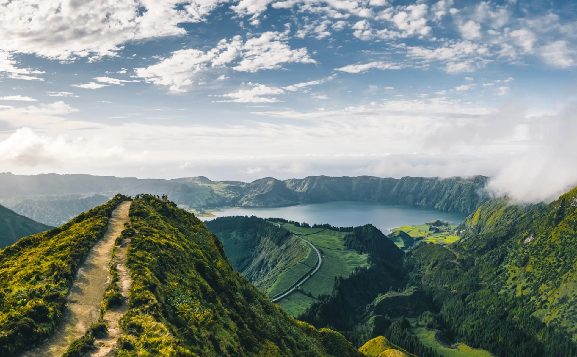 A scenic mountain path leading to a lake surrounded by lush green hills under a partly cloudy sky.