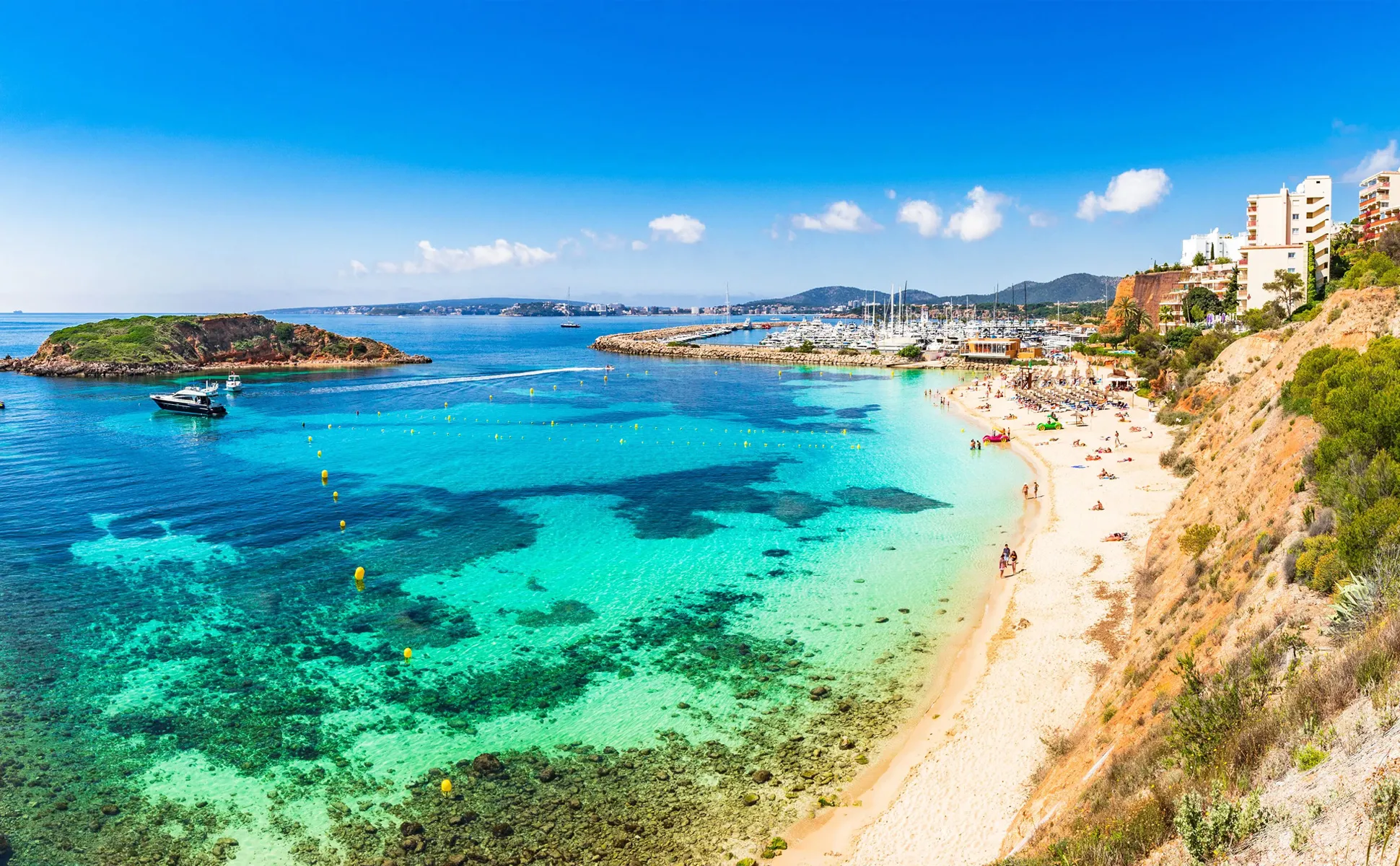 Clear turquoise sea and sandy beach at a coastal town with boats and buildings near the shore.