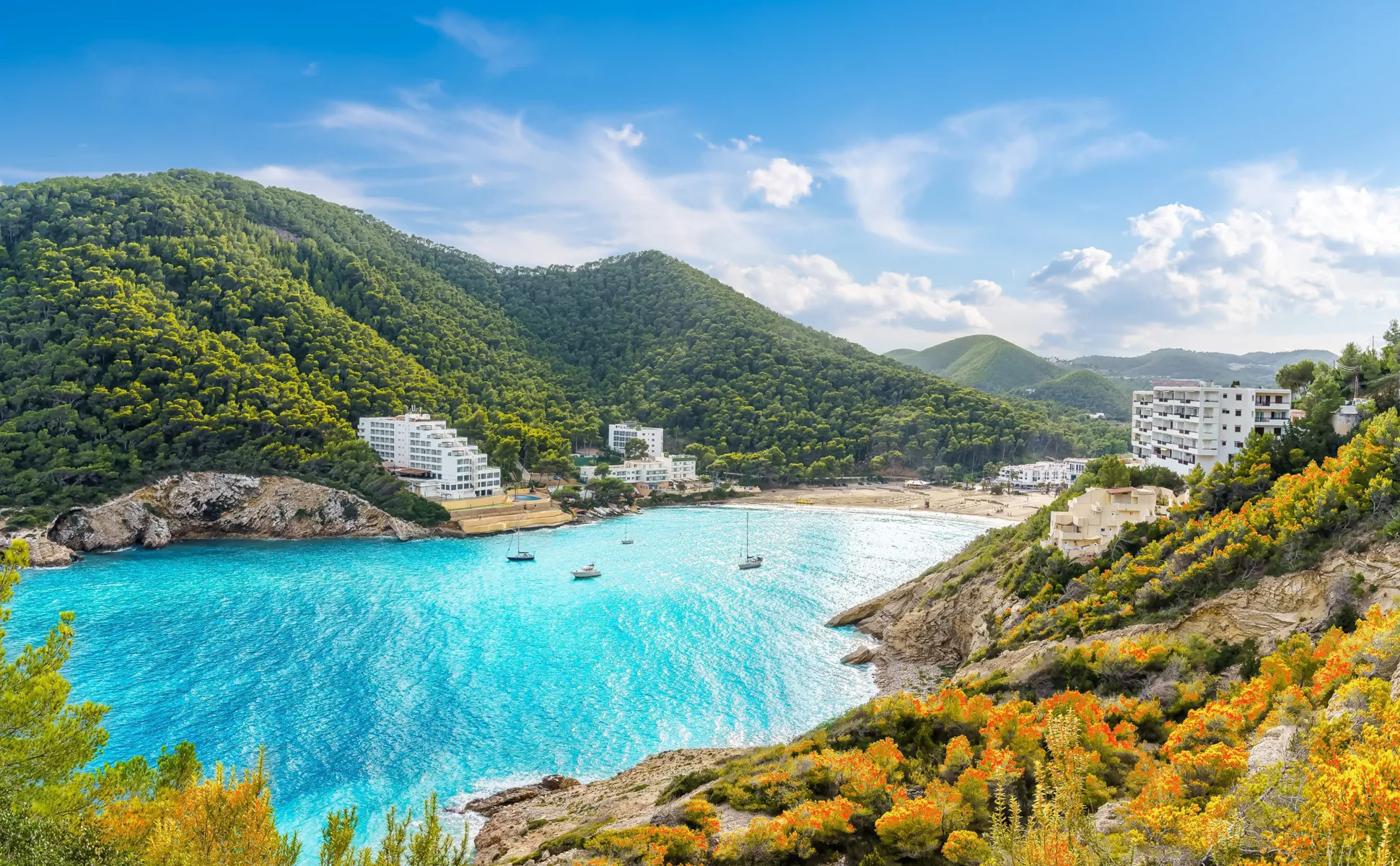 Coastal bay with turquoise water, boats, green forested hills, and white buildings near the beach for car rental visitors.