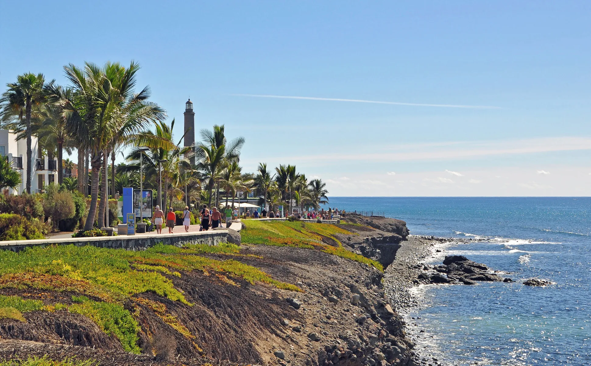 Coastal promenade with palm trees, people walking, rocky shore, and lighthouse by the ocean.