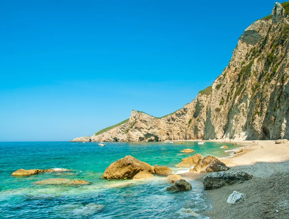 Spiaggia di Assos a Cefalonia con acque turchesi, scogliere rocciose e cielo soleggiato.