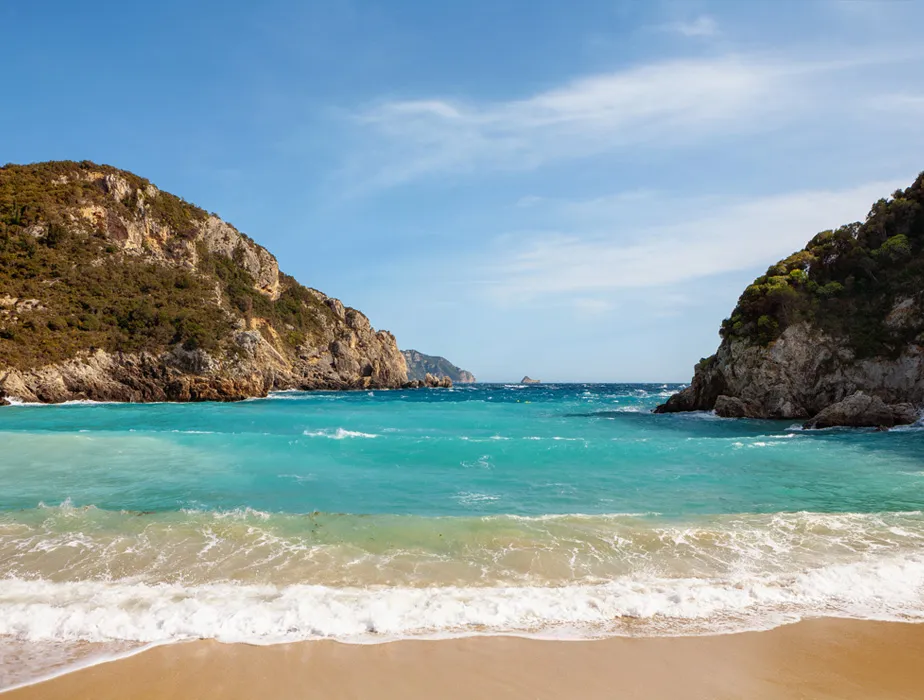 Spiaggia sabbiosa isolata con acqua turchese chiara tra scogliere rocciose sotto un cielo parzialmente nuvoloso.