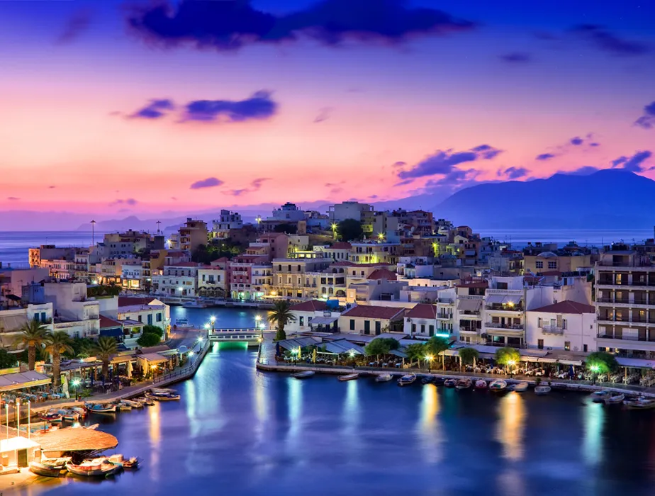 Agios Nikolaos town and harbor on Crete at sunset with calm waters and illuminated buildings for car rental.