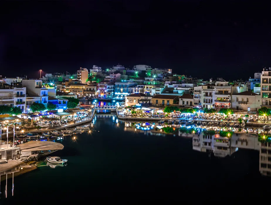 Agios Nikolaos harbor in Crete glowing with lights and reflections at night near car rental spots.