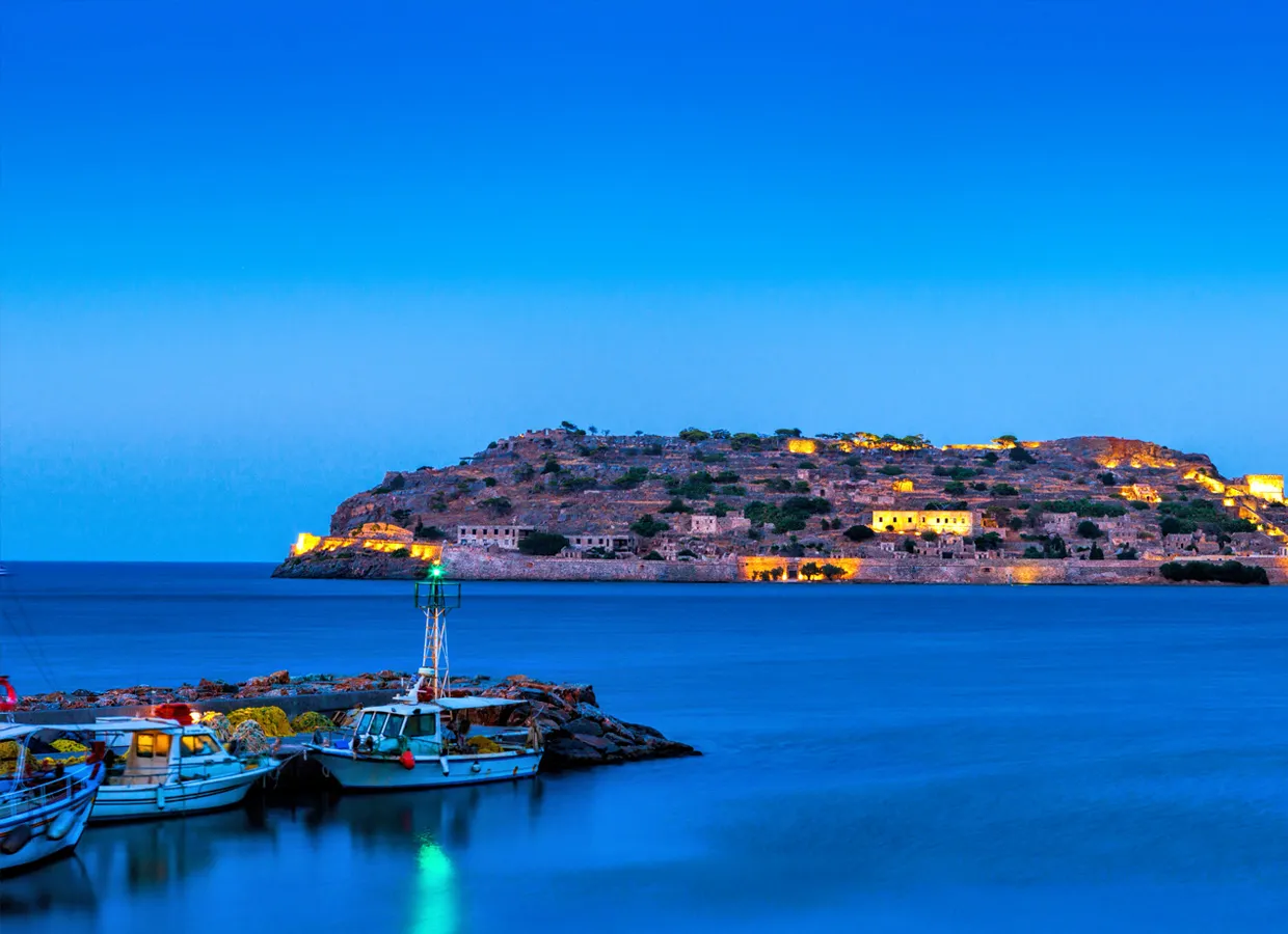 Spinalonga Island in Greece at dusk with boats moored in calm blue waters and illuminated ruins visible.