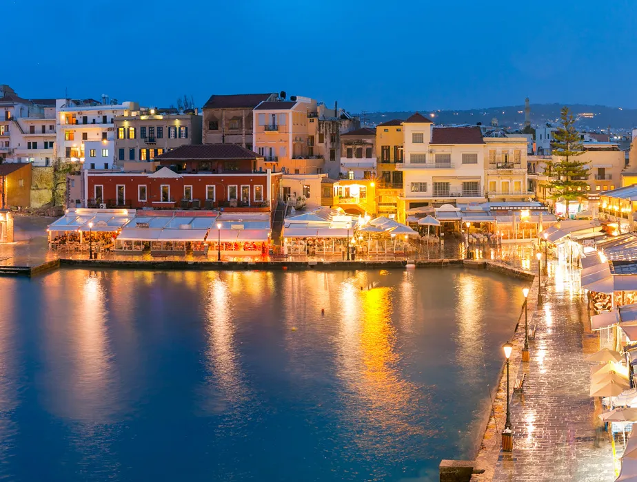 Chania old harbor lit up at night with waterfront restaurants and calm sea reflections for car rental.