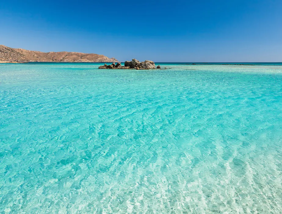 Clear turquoise water at a shallow beach with rocky hills in the background under a blue sky.
