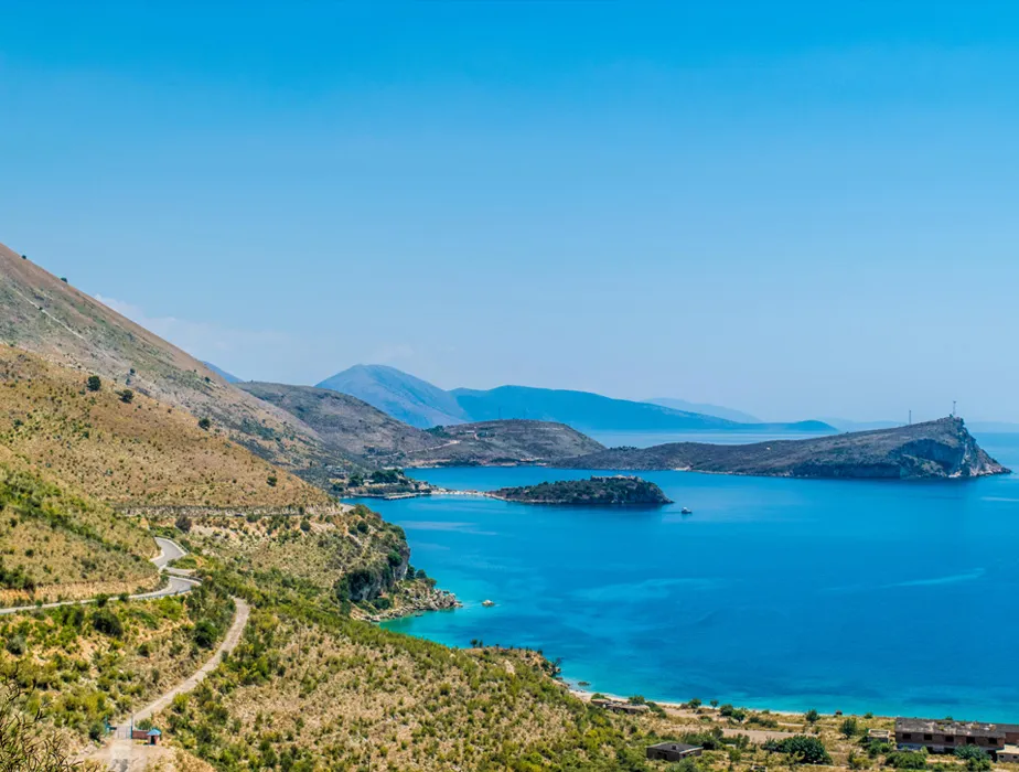 Coastal road winding along clear blue sea and hills under a bright sky in a Mediterranean location.