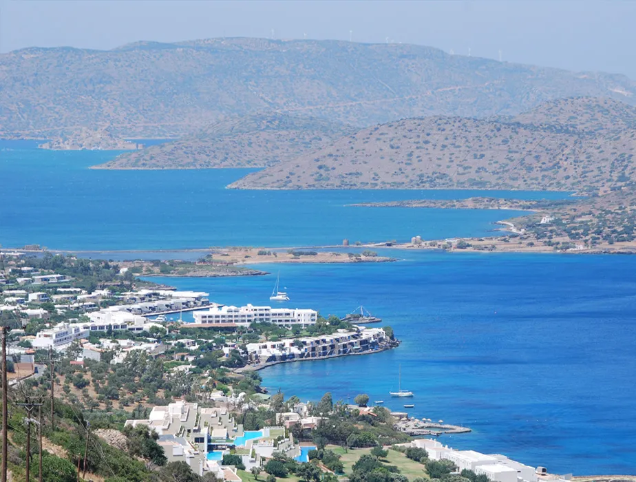 Elounda coastline with blue sea, boats, and white buildings, ideal for car rental travel.