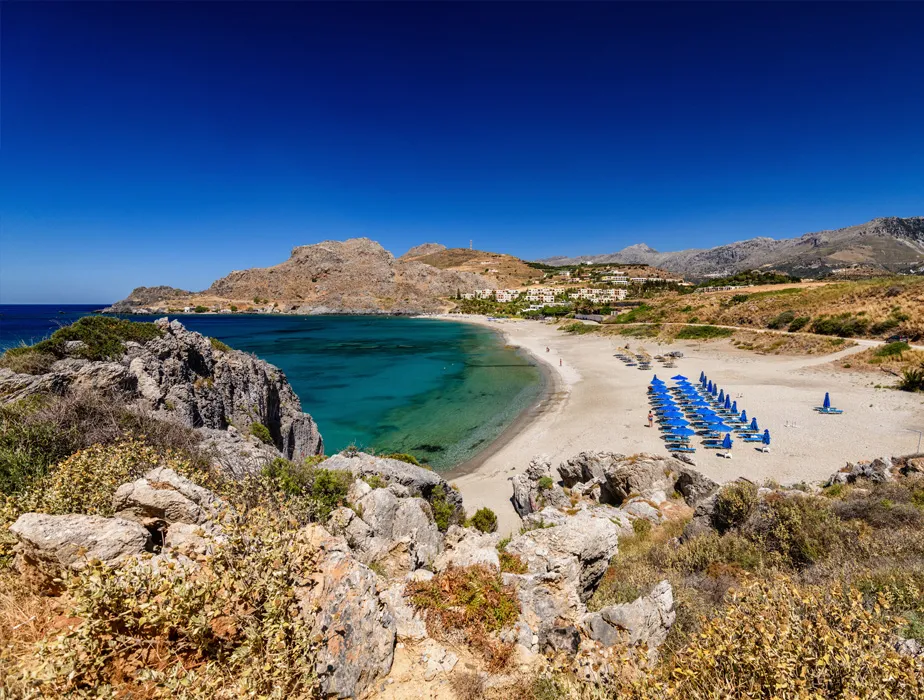 Lindos beach with clear blue water, sandy shore, and blue umbrellas, Rhodes island, Greece car rental spot.