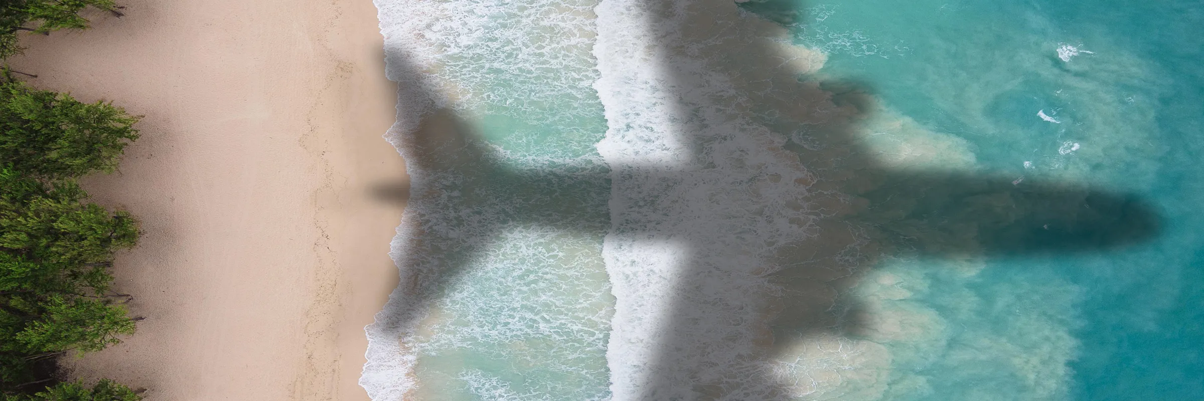Schatten eines Flugzeugs über einem Strand mit türkisfarbenem Wasser und grünen Bäumen am Ufer.