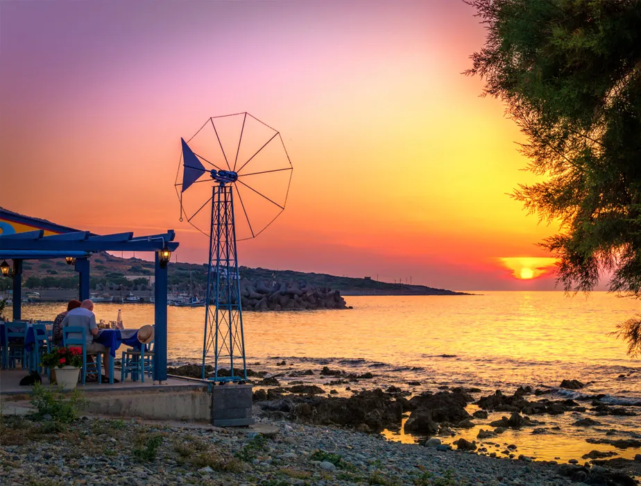 Seaside dining at sunset with a traditional windmill near the coast in a Greek island setting.
