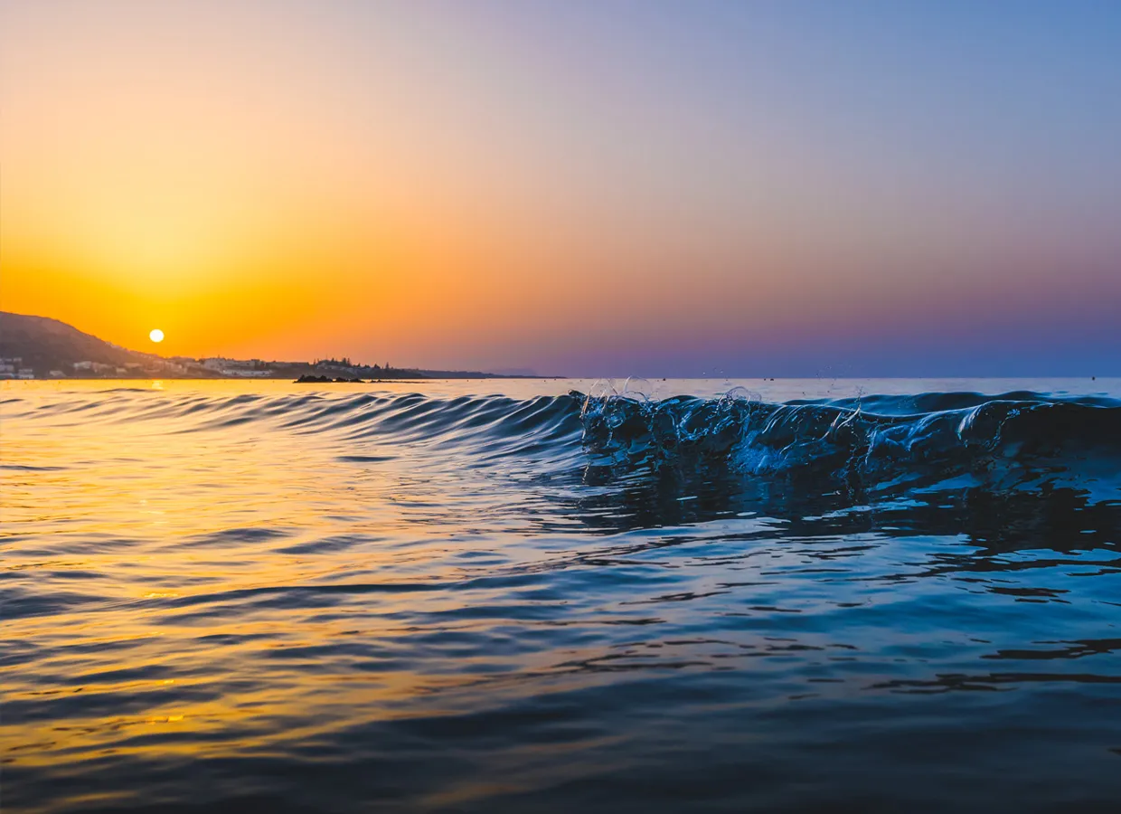 Sun setting over calm sea waves with a clear sky and hills in the distance by the shore.